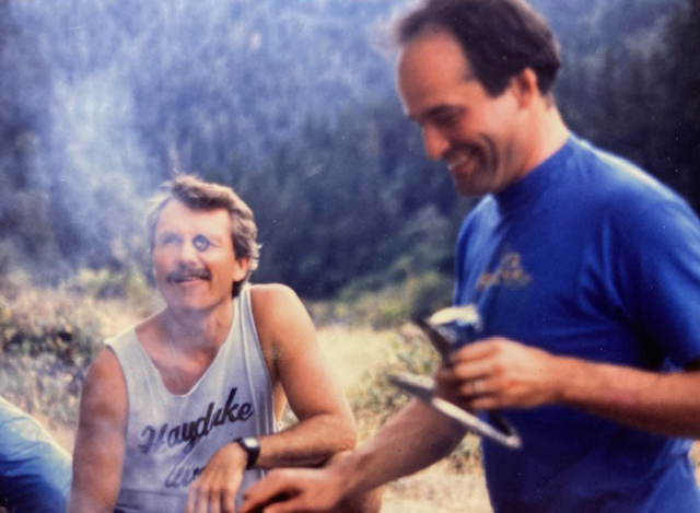 Dr. John Kitzhaber and Bill Bradley camping on Rogue River. Dr. Kitzhaber is holding a Pickapeppa bottle cap in his eye and Bil Bradley is cooking.